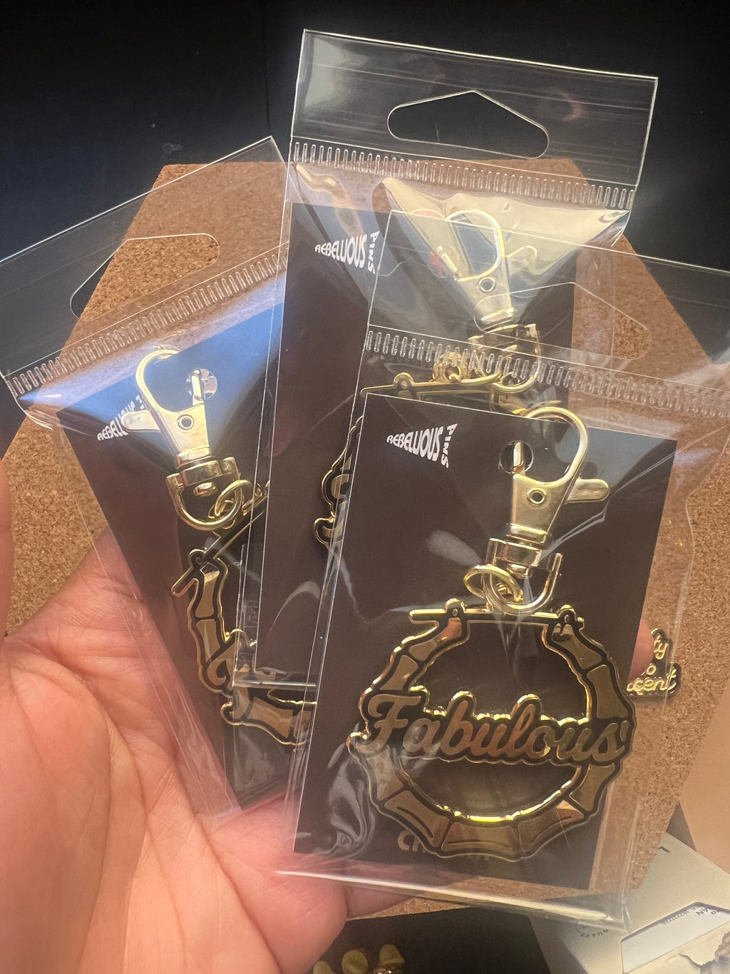 Gold 'Fabulous' keychain in clear packaging held by a hand.