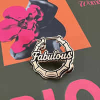 A close-up of a book or magazine cover titled 'BLOOM' with a dark olive-green background, a large pink flower illustration, and the word 'Women' near the top right. A decorative metal pin reading 'Fabulous' is placed over the lower center of the cover.