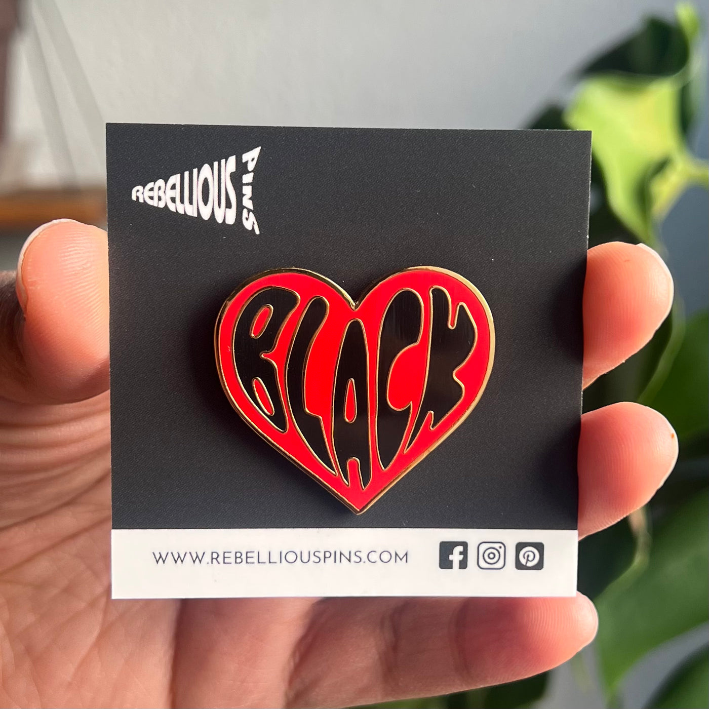 A hand holding a hard enamel pin with a heart shape and the word 'BLACK' in bold letters.