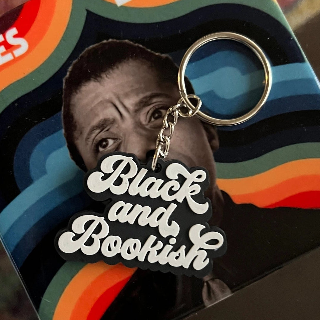 A James Baldwin 'Black and Bookish' keychain in its original square packaging, displayed on a dark reflective surface with a patterned rug in the background.
