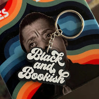 A James Baldwin 'Black and Bookish' keychain in its original square packaging, displayed on a dark reflective surface with a patterned rug in the background.