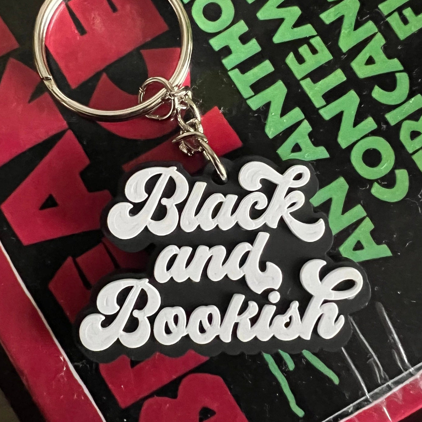 A close-up of a keychain with a silver metal split ring and charm that reads 'Black and Bookish' in white script, placed on top of a book cover with bold red and green text.