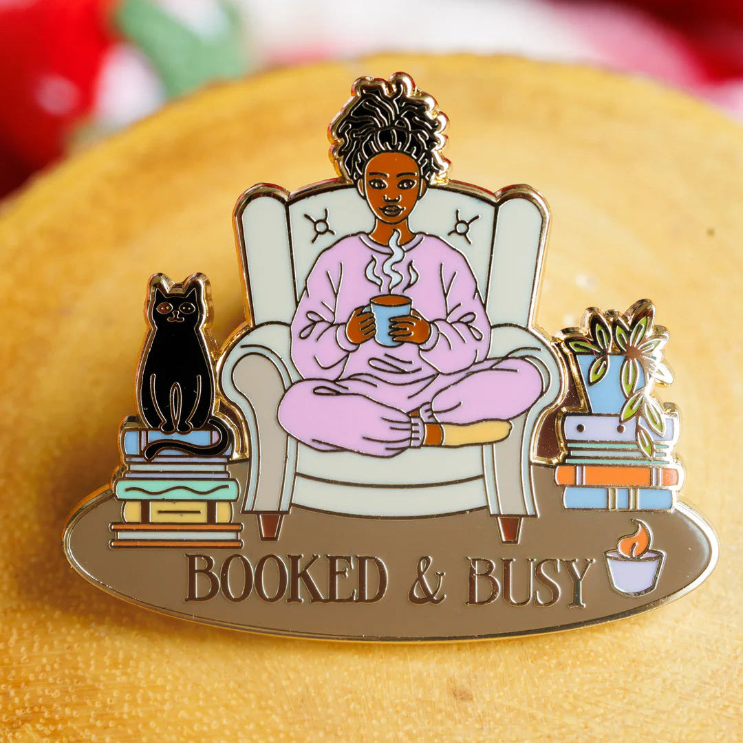 Enamel pin of a person sitting in a chair with a cat, holding a mug, and the text 'Booked & Busy' on a wooden surface.