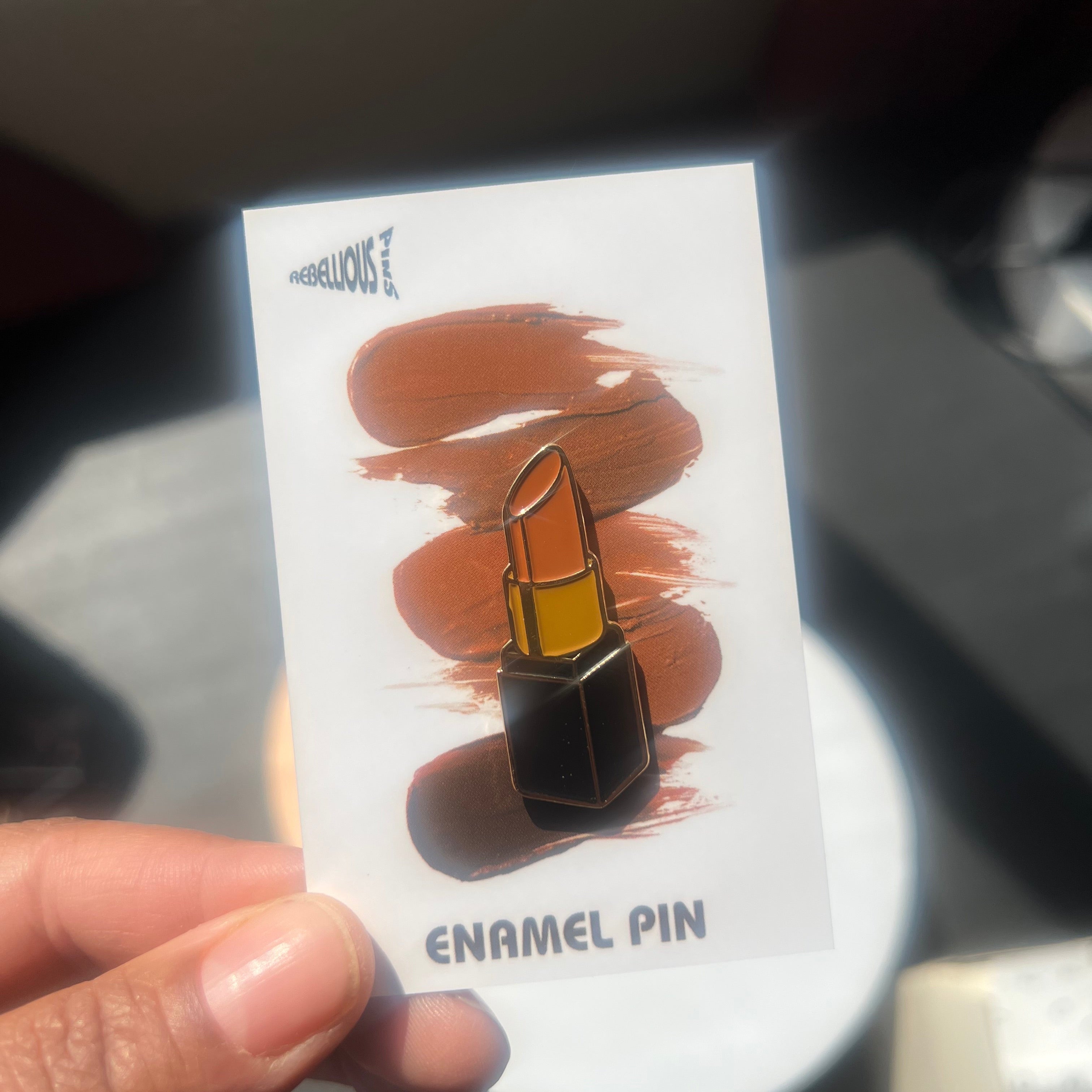 Lipstick Enamel pin on a card held by a hand with a blurred background