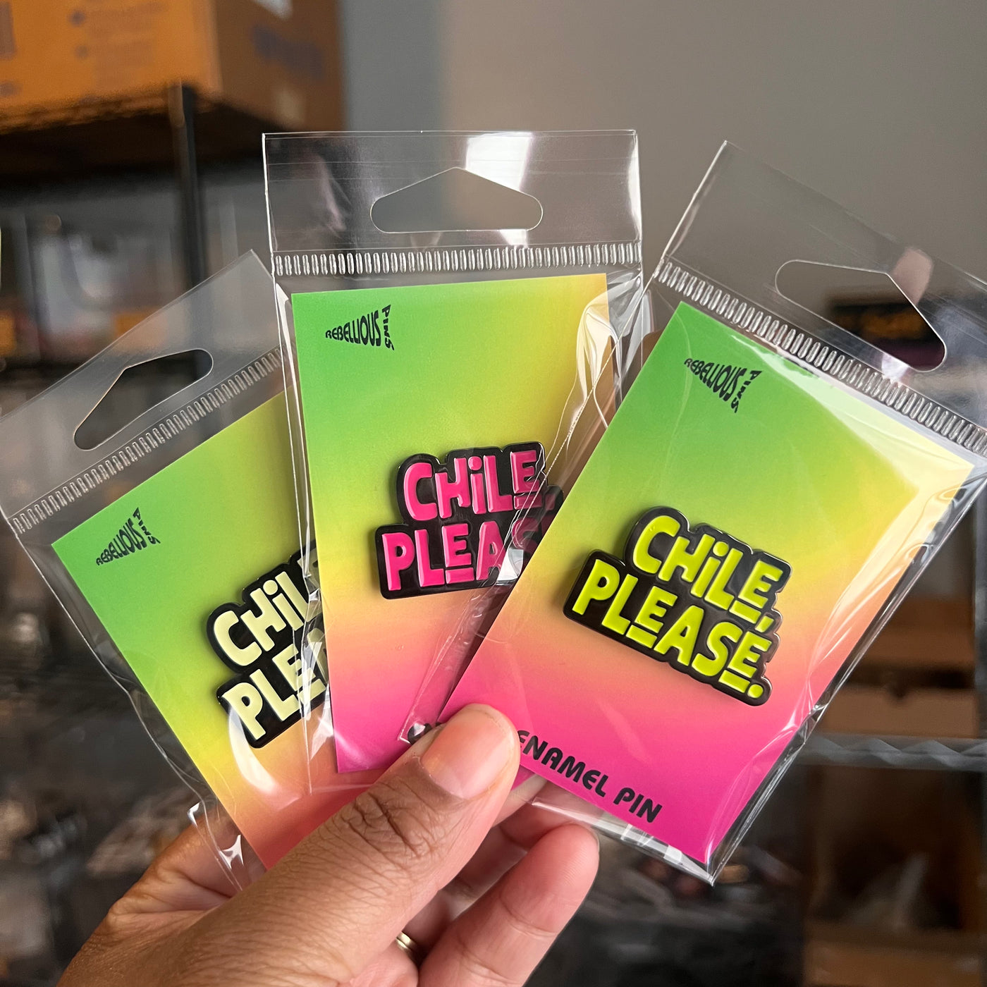 Three 'Chile Please' enamel pins in clear packaging held by a hand.