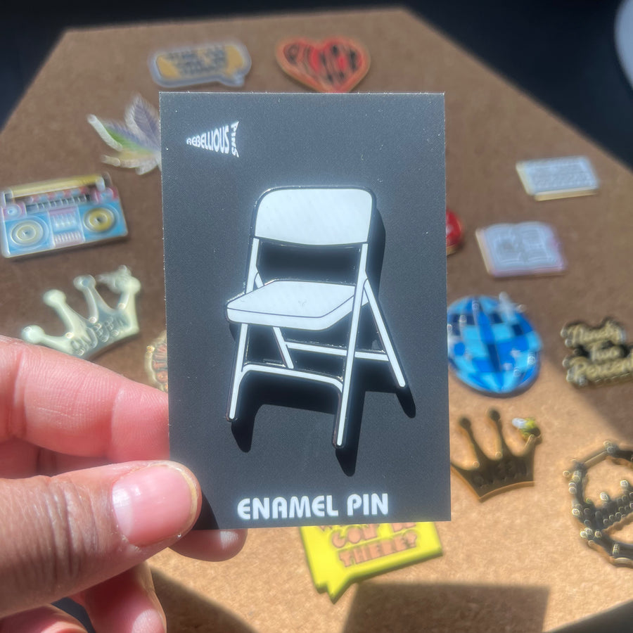 Enamel pin card with a chair design held by a hand, with various other pins in the background.
