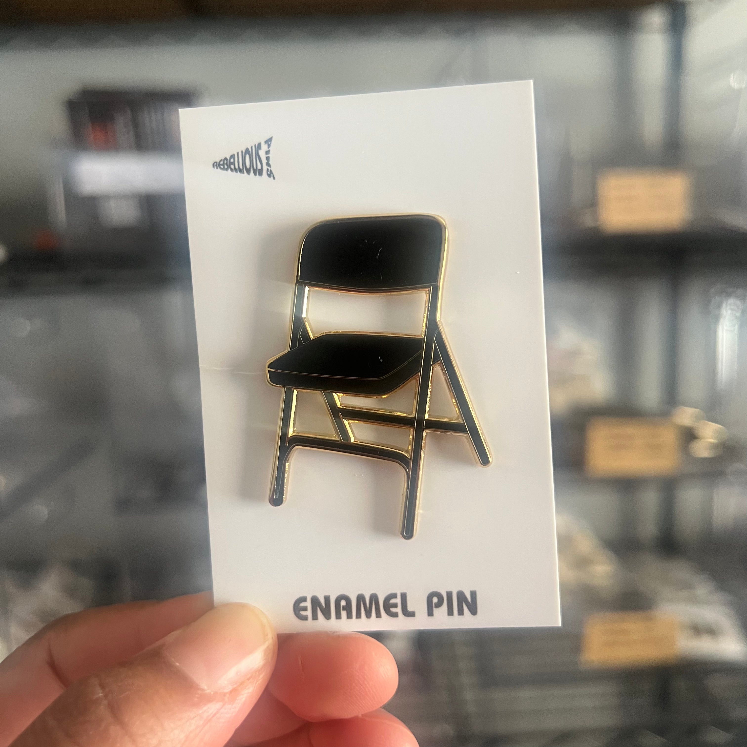 Enamel pin of a black chair on a white card held in front of a blurred background