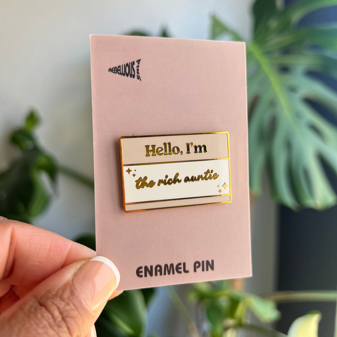 Enamel pin on a pink card held by a hand with a plant in the background
