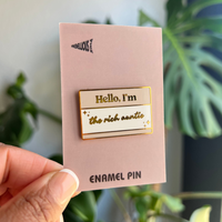Enamel pin on a pink card held by a hand with a plant in the background