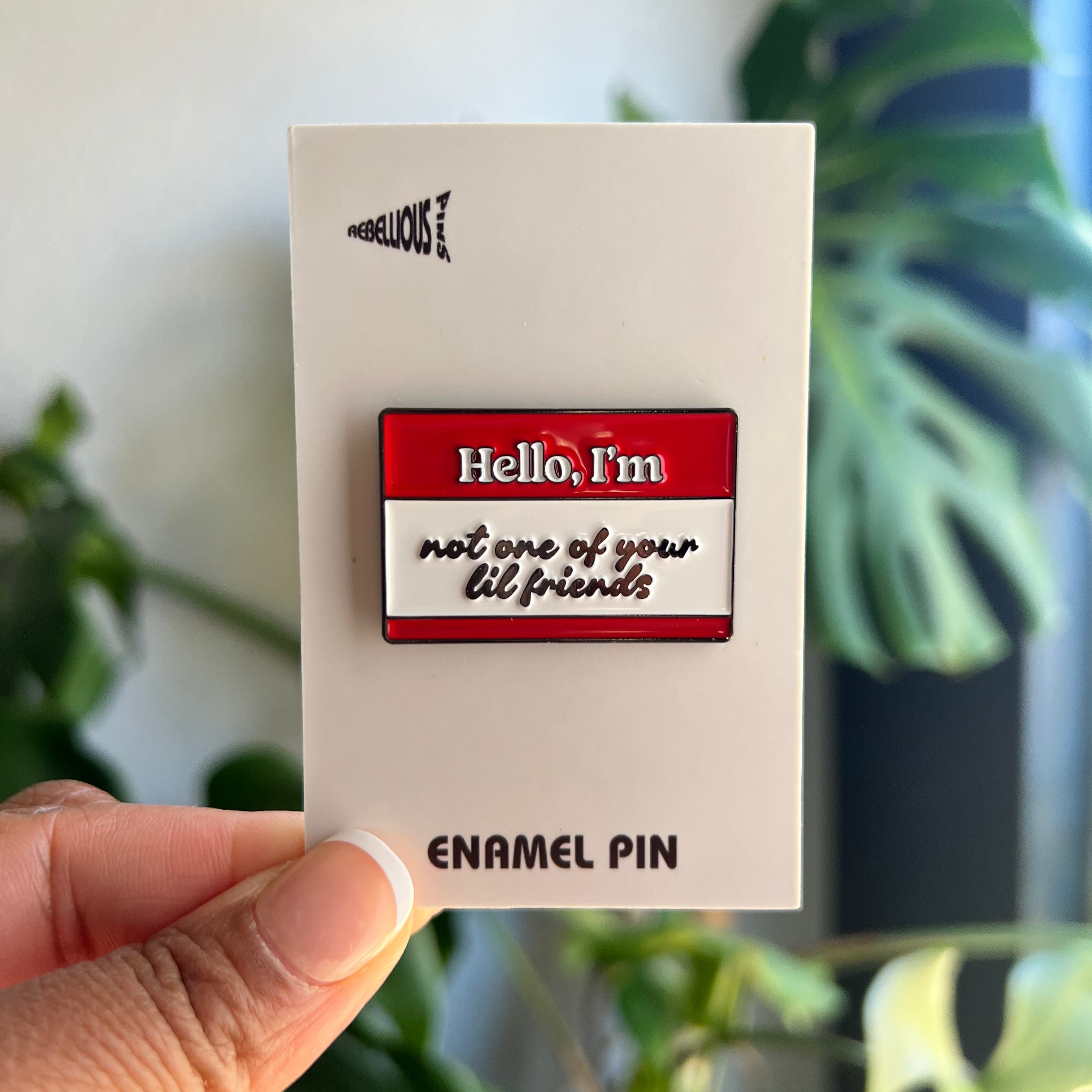 Enamel pin with 'Hello, I'm not one of your lil friends' text on a card held by a hand with plants in the background.