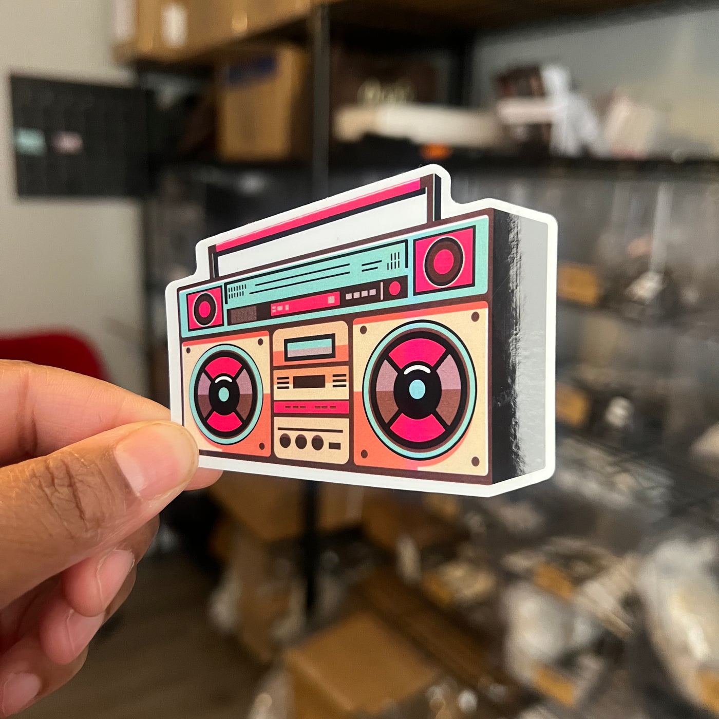 Sticker of a retro boombox held by a hand in an indoor setting.