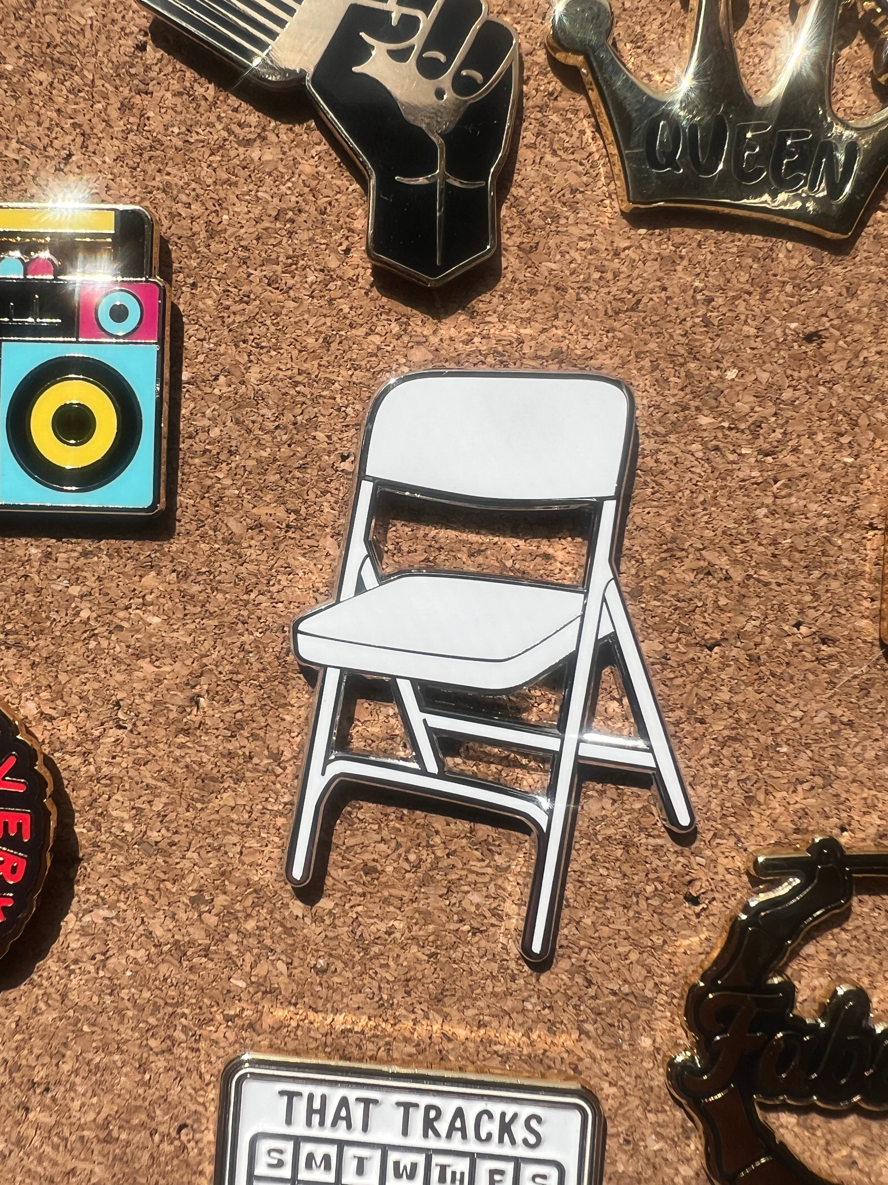 Pin with a white folding chair design on a cork board with other pins.