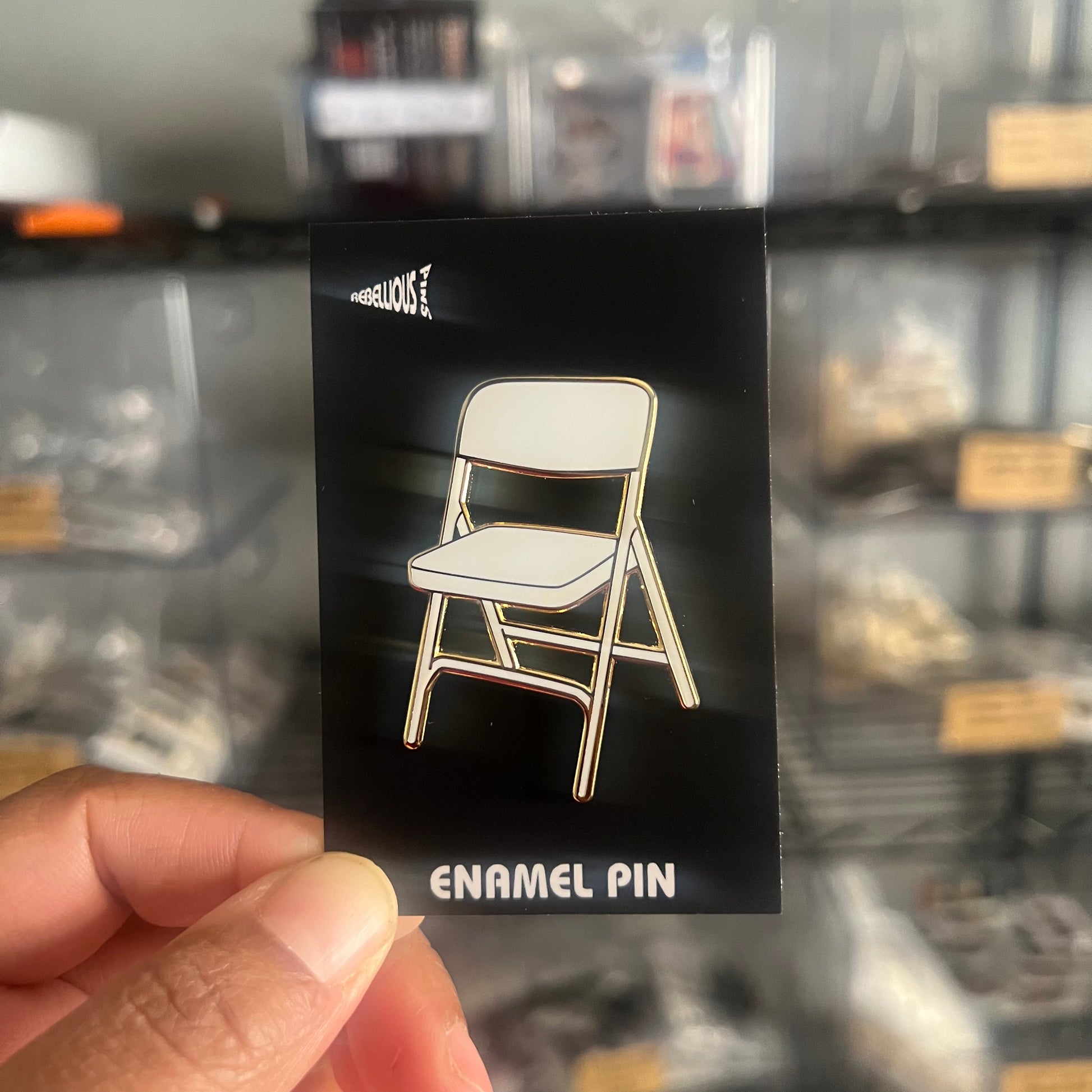Enamel pin of a chair on a black card held by a hand with a blurred background.