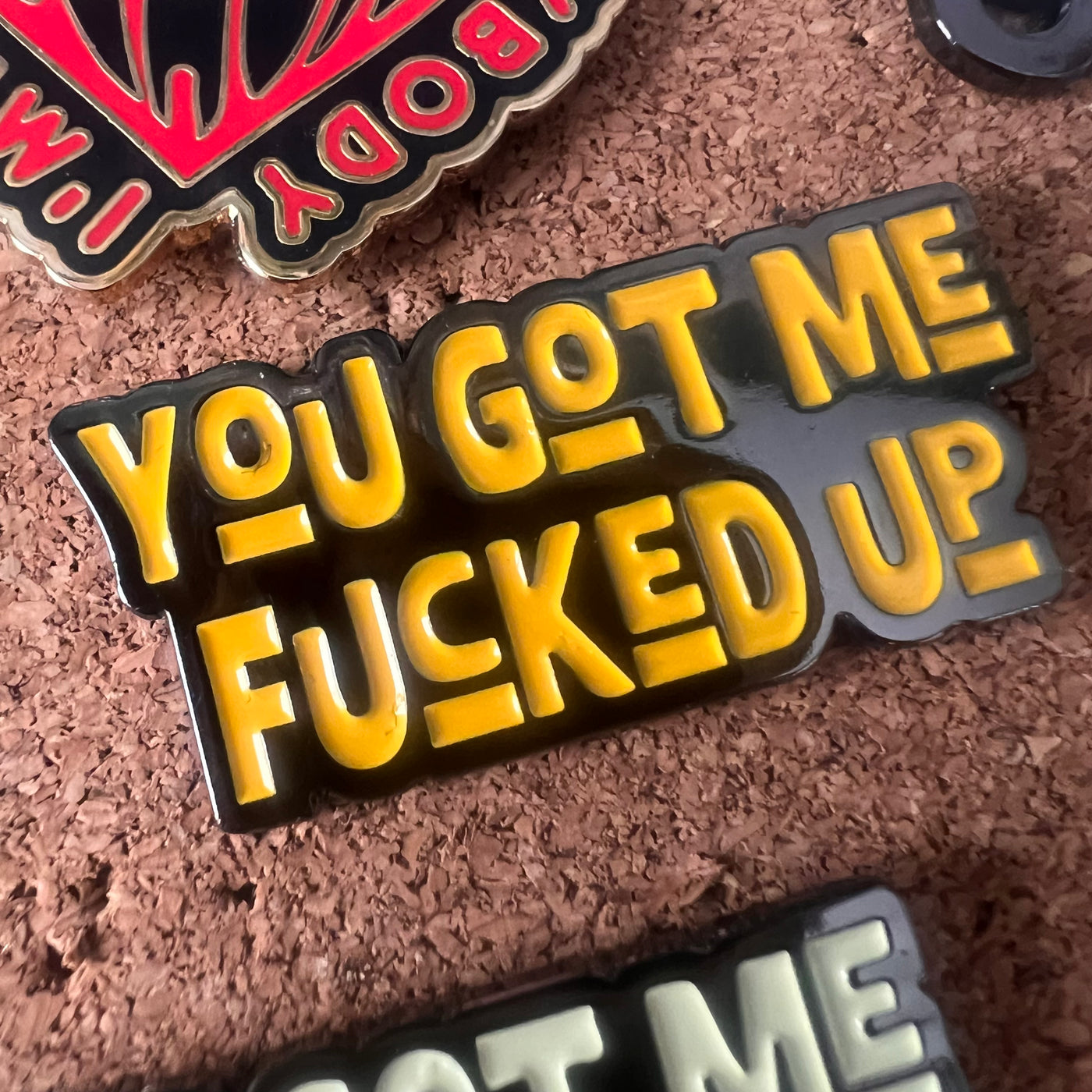 Enamel pin with yellow text 'YOU GOT ME FUCKED UP' on a cork background