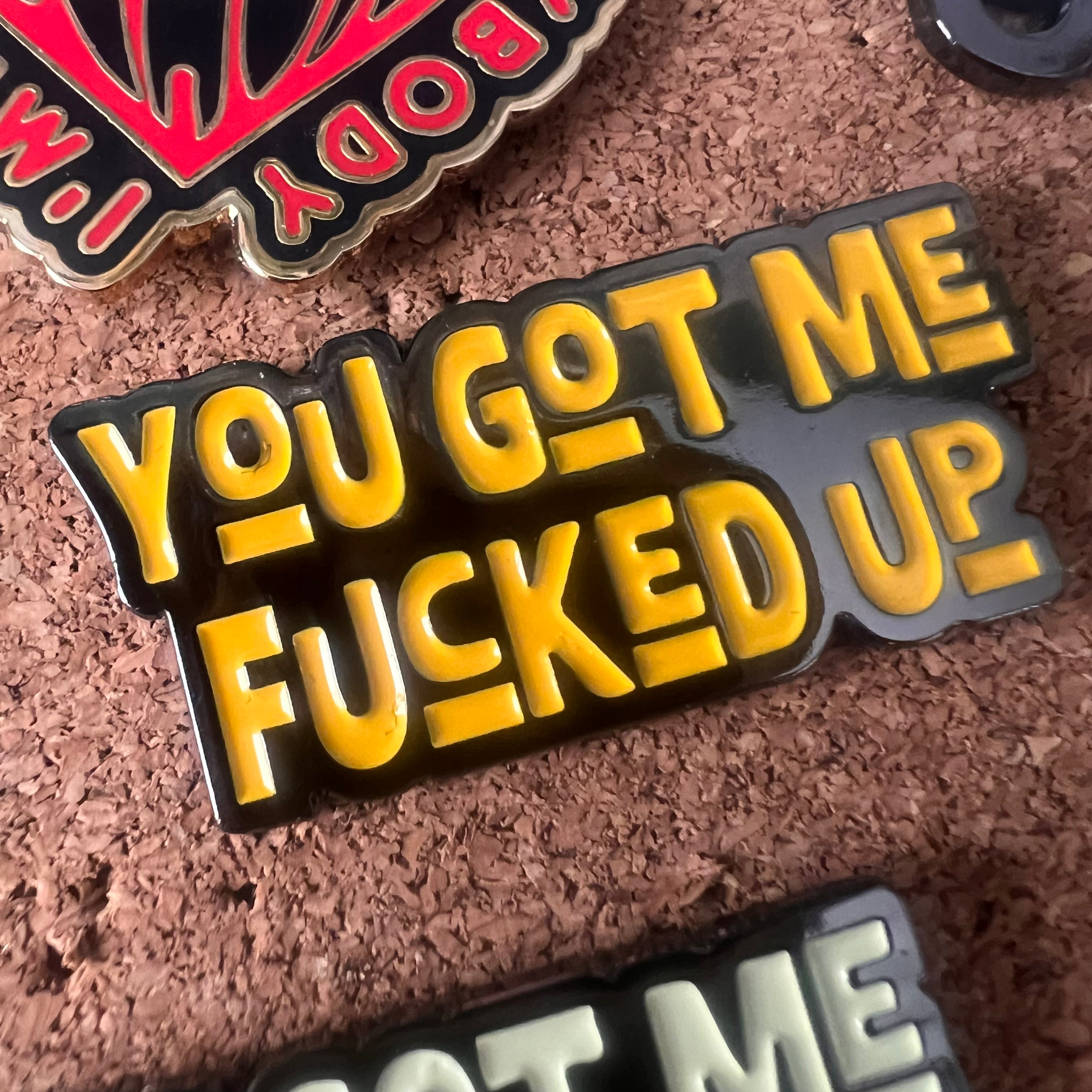 Enamel pin with yellow text 'YOU GOT ME FUCKED UP' on a cork background