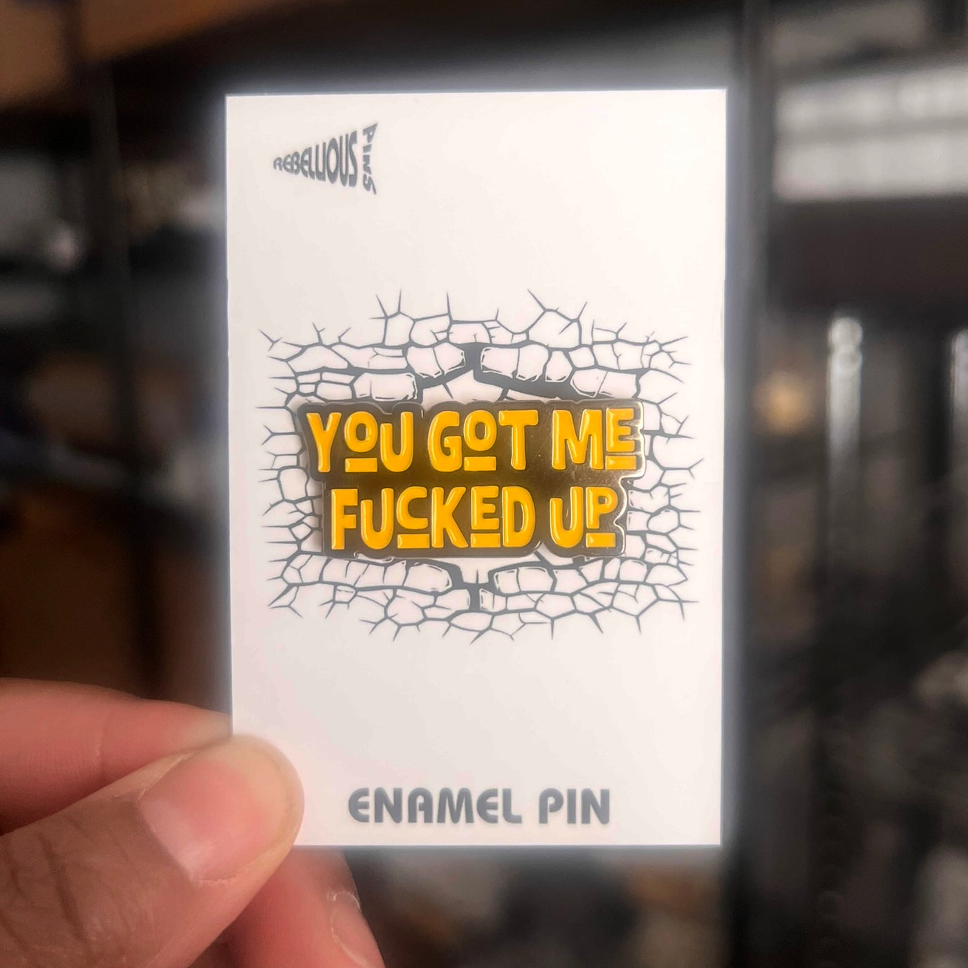 Enamel pin with cracked design and text held by a hand against a blurred background