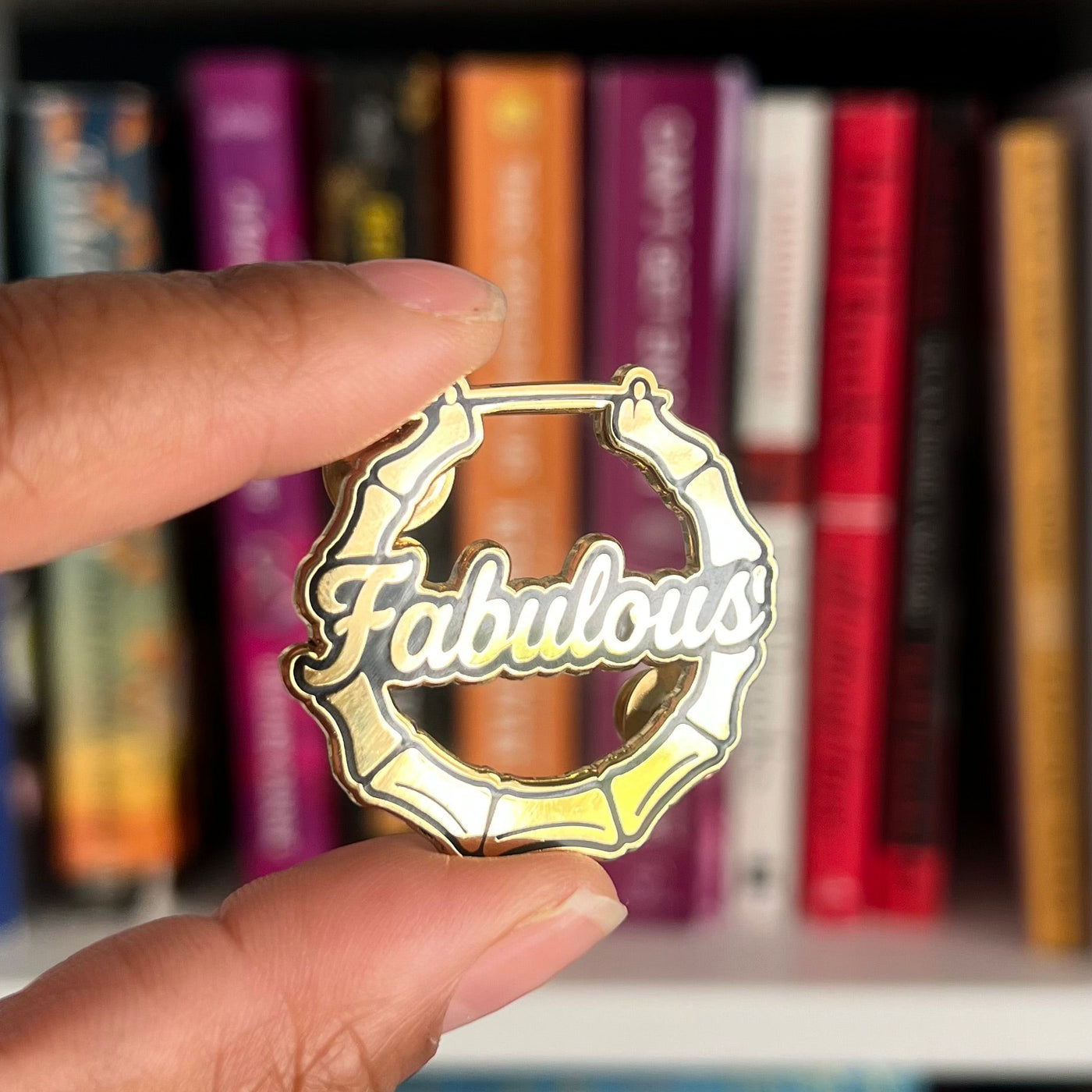 A hand holding a circular enamel pin that reads 'Fabulous' in white script over a gold and yellow geometric frame, with blurred bookshelves in the background.