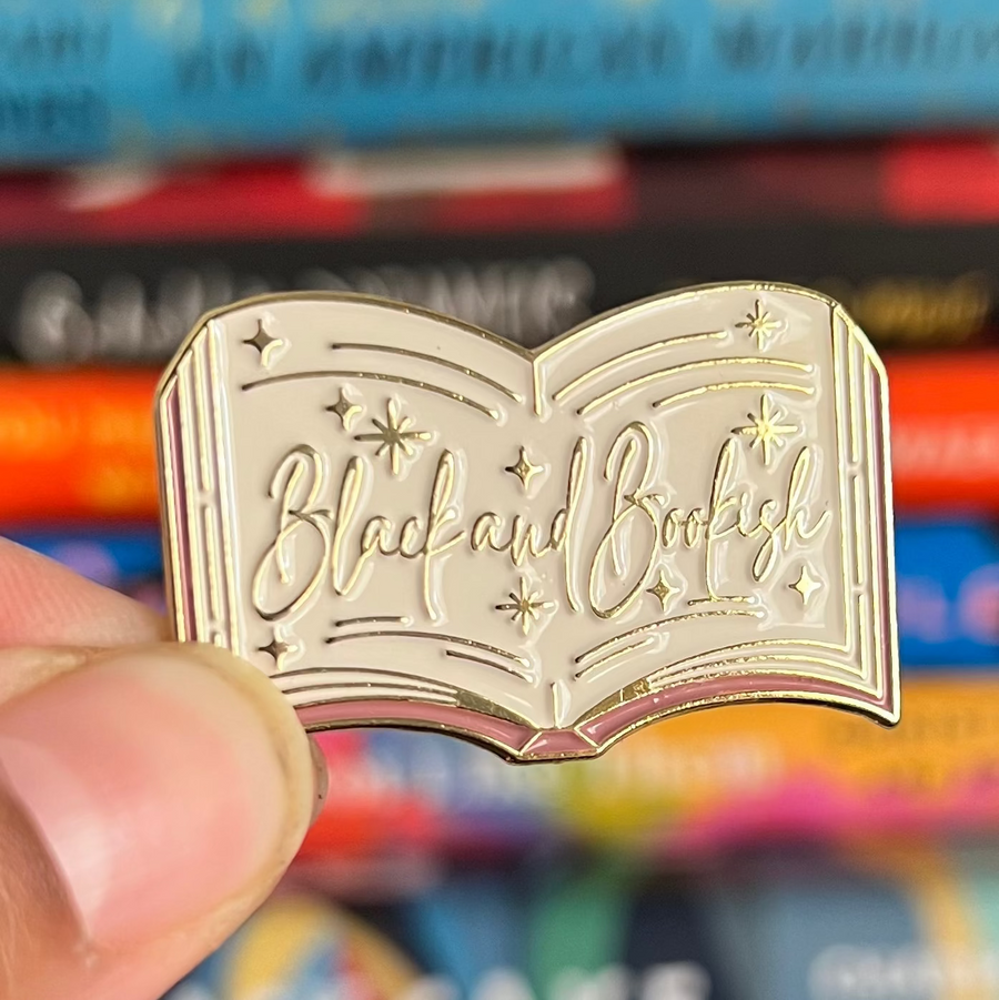 Gold-colored 'Black and Bookish' book-shaped pin held in front of a stack of books.