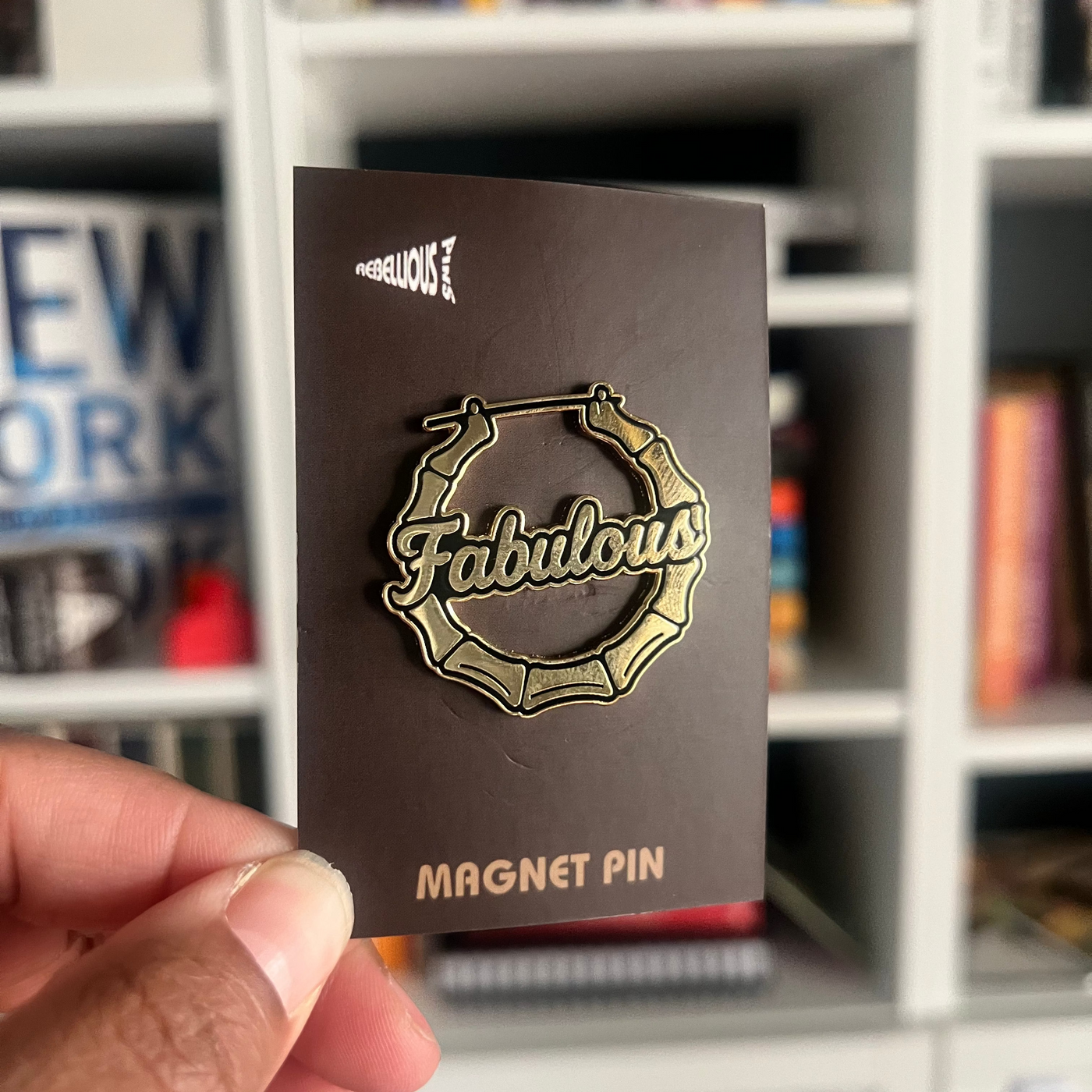 Magnet pin with 'Fabulous' design held in front of a bookshelf.