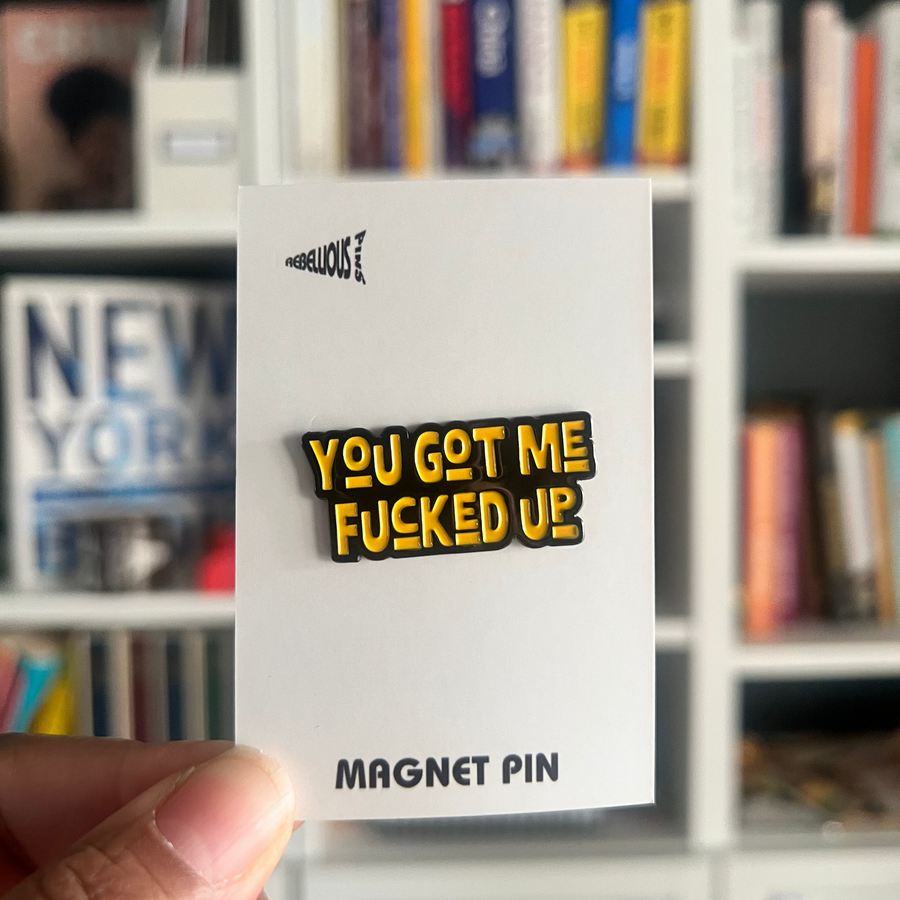You Got Me Fucked Up Magnet Enamel Pin on a backing card pictured in front of a bookshelf.